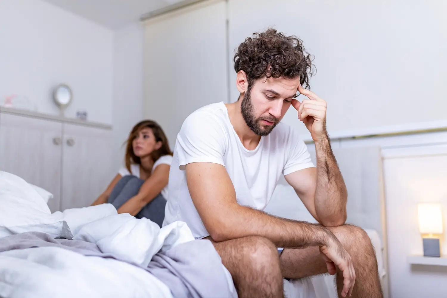 Concerned man sitting on bed with a woman in the background.